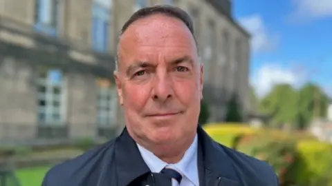 A man with short grey hair looks at the camera in a close-up shot. He is wearing a light blue shirt and dark blue jacket and tie. He is standing in front of a stone building and a garden.