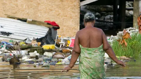 A woman wrapped in a green patterned sarong wades through knee-deep water to reach her home. 