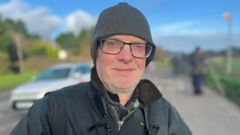 Man wearing a grey woolly hat, with binoculars around his neck. He has black rimmed glasses