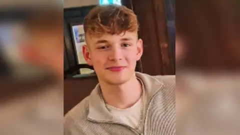 George, a teenager with curly blond hair shaved at the sides and long at the top, smiles into the camera while wearing a beige coloured fleece open at the neck.