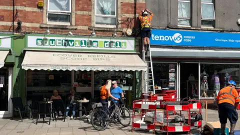 A workman works up a ladder on electrical infrastructure on the front of shop buildings. There are more workmen on the street and pedestrians walking by