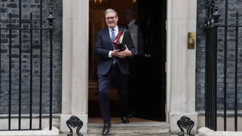 Lucy North/PA Wire Sir Keir Starmer leaving 10 Downing Street ahead of PMQs - he is carrying folders and is dressed in a navy suit with a blue tie. He has grey hair and is wearing dark-rimmed glasses.
