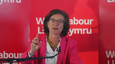 Eluned Morgan speaking in front of a red Welsh Labour banner, wearing a pink jacket. She has a white shirt underneath with a gold chunky necklace on. She has purple framed glasses and dark brown shoulder-length hair. 
