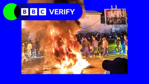 Huge fire and plume of smoke in the centre of the image with crowds of masked people surrounding it during Tehran protest 