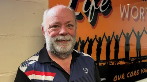 Martin Fletcher stands in front of an orange poster. Wearing a dark blue sports top.