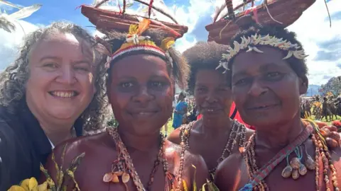 Nancy Irwin A group of four people pose closely together outdoors during what appears to be a cultural gathering or community event. Several people are wearing traditional adornments, including shell necklaces and decorative headpieces, with woven items balanced on their heads. The scene is brightly lit by sunlight, with a grassy field, other attendees, and a partly cloudy sky visible in the background.