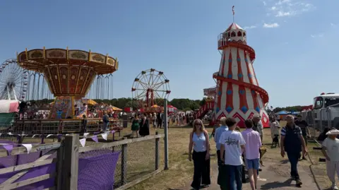 Liz Nice/BBC A red white and blue helter skelter stands next to a carousel with a big wheel and blue sky in the background
