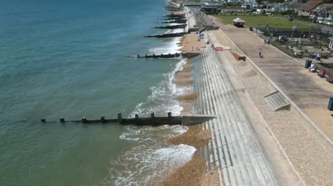 An aerial shot of a shingle beach with wooden groynes placed along it. Behind the beach is a concrete promenade and a square of grassland. Some houses lie beyond it.