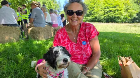 Jennie Brown with her dog sitting on a grass bank. She is wearing a pink top and black sunglasses.