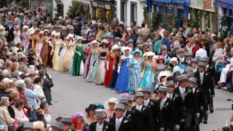 Helston Flora Day Association About 30 couples are walking along a street which is lined with spectators. The ladies are wearing brightly-coloured full-length gowns with hats and gloves. The gentlemen are wearing top hats and tails. The shops lining the street are decorated with greenery and flowers.