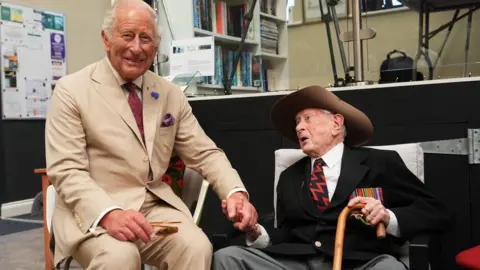 PA Media The King smiling while sitting in a chair next to James Fenton, whop is holding his hand.