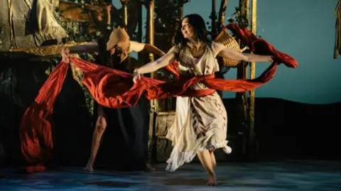 supplied A woman, dressed in a white flowing dress and billowing red sash, dances on stage.
