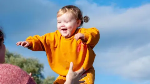 A little girl in an orange jumper and trousers is playing with her mum outside on a sunny day. The mum has her back to the camera and is opening her arms to catch the girl. 