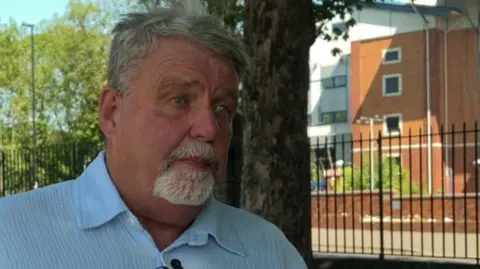 A man with short grey hair and a beard wearing a blue shirt.