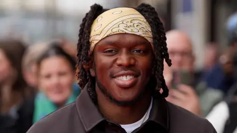 KSI smiles and wears a light yellow bandana over his hair with a white t-shirt and black shirt on top.