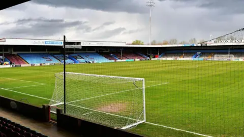 A view of the pitch at Glanford Park football stadium from behind a goal. The grass is surrounded by blue and claret-coloured seats. A floodlight can be seen at one end of the ground.