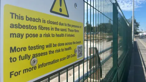 A picture of a sign which is yellow and white in colour. It is on a large green fence.