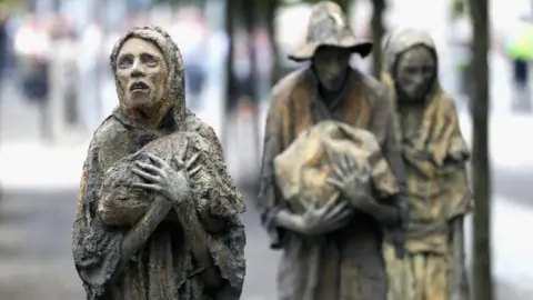 Getty Images Bronze sculptures, three emaciated humans, gripping bags. 