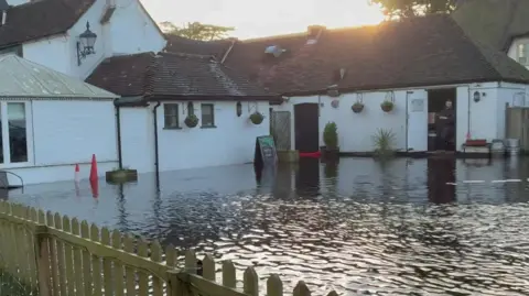 The Woolpack pub, which has white walls and a red tiled roof, can be seen surrounded by floodwaters. 