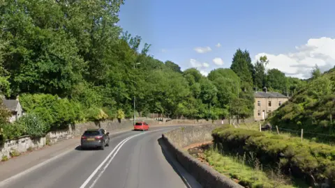 A country toad with head-height stone walls either side. Beyond the walls are tall trees on the left hand side, and a stream on the right.