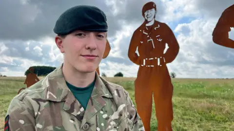 Rifleman Izzy O'Malley in uniform, standing in front of the statue modelled on her.