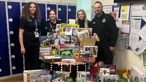 East of England Ambulance staff standing behind a large selection of toys. They are all wearing a dark uniform with two with a blue landyard round their necks.
They are Olivia Howard, Sophie Bailey, Georgina Humphreys and Stephen Notley