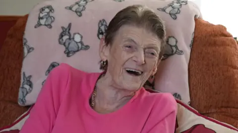 Shaun Whitmore/BBC An older woman is laughing while sat in a large chair. She is wearing a pink top.