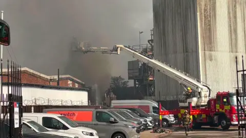 A fire appliance is parked at the scene of a bathroom showroom fire. An aerial ladder is being used to tackle the blaze which a firefighter is on, however, it is hard to see them among a dark plume of smoke.