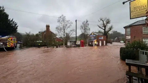 Dark orange floodwater running through a village. The water has covered the road and is sweeping through the street. There is a pub on the right and a bus stop, phone box and fish and chip shop that are all flooded. A fire engine is on the left trying to pump out the water.