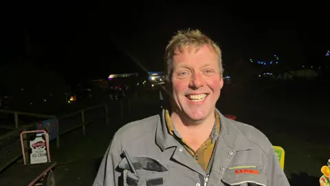 John Devine/BBC Andy Johnson is smiling. He has short sandy coloured hair and is wearing a light green boiler suit with a mustard tartan shirt underneath, the picture is taken at night and there are fairy lights visible in the background.