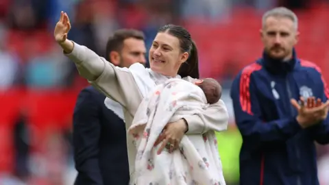 Hannah Blundell smiles and waves while holding her baby on a football pitch