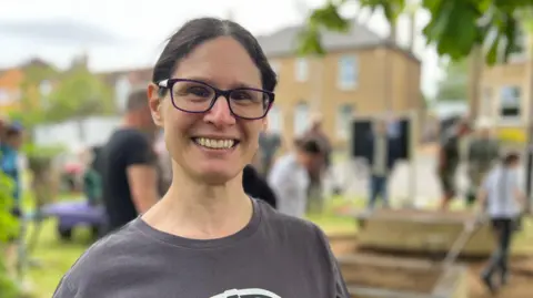 Tom Jackson/BBC Lisa Burton is standing on the left of the frame on a small patch of grass which is surrounded by houses. Burton has brown hair in a low ponytail and is wearing black-framed glasses and a dark grey T-shirt. There are several people standing in the background and are blurred.