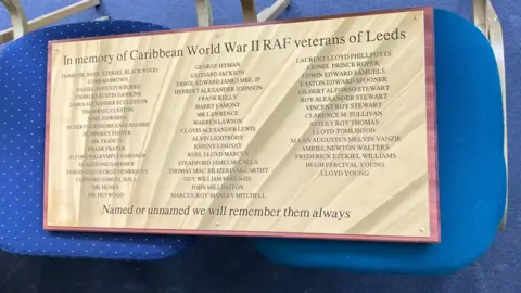 A gold plaque inscribed with the names of 49 veterans in a list format is mounted on wood, laid across two blue chairs.