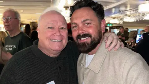 Michael Starke, 68, who as a bald head and wears a dark brown jumper, smiles and poses with his arm around the shoulders of Ray Quinn, who has dark hair swept back and a dark beard and wears a beige woollen jacket.