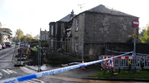 PA Media A grey sandstone block containing several properties. There is police tape strung along to a no entry road sign, and metal fencing cordoning off the property. People in protective clothing are working in the front garden. There is a large hole on the upper floor and the roof, and debris from inside the flat can be seen sticking out of the gap.