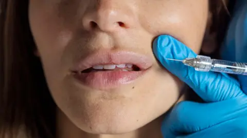 A stock image of a woman with only the lower half of her face visible about to receive an injection with her mouth being pulled upwards slightly by a blue-gloved hand.