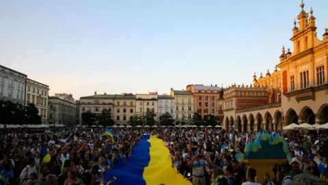 Getty Images A large crowd of people in a square surrounded by buildings on three sides. A large blue and yellow Ukrainian flag is displayed running through the centre of the crowd. 24 August 2023