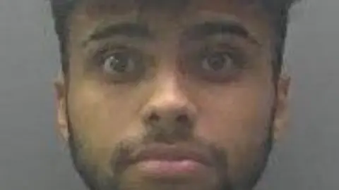 Cambridgeshire Police Mugshot of Isaiah Khan. He is brown and has dark hair [ short back and sides and long on top]. He has a dark beard and moustache, which is groomed close to his skin. 