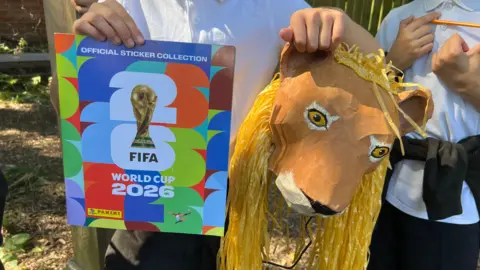 Jessica Bayley/BBC Image shows a close up of a FIFA World Cup 2026 sticker book which is multicoloured and has a picture of the World Cup on the front. It also shows a paper mache lion head.