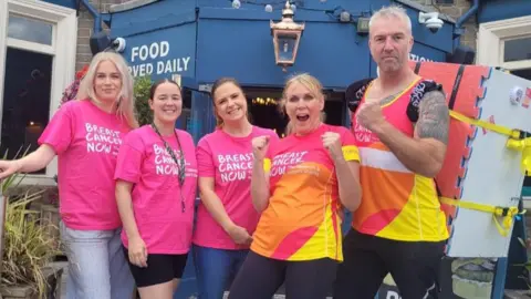 A man and a woman dressed in brightly coloured matching sports gear hold up their arms in celebration next to three women dressed in pink t-shirts
