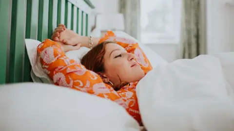 A woman lying in bed looking content after a great night's sleep. She is wearing orange pyjamas in a bed with white sheets and green headboard