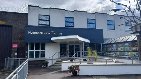 Exterior shot of Hydebank Wood College, a white building with navy signage and colourful art on the wall. There is barbed wire across the roof of the building.