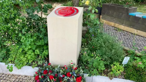 BBC/Adele Forrest The plinth sits in a flowered garden surrounded by red wreaths