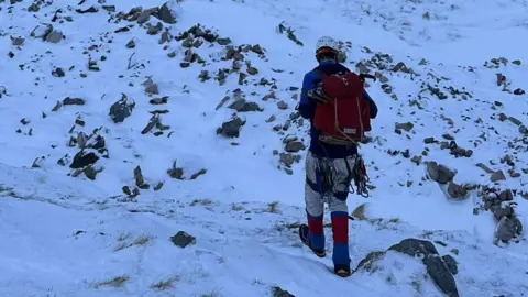 Brian McGillicuddy, photographed in the snow from behind. He is wearing a white helmet, blue jacket, red rucksack, white trousers, red and blue gaiters and a climbing harness with gear.