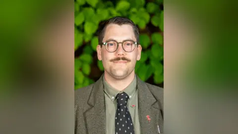 Welwyn Hatfield Borough Council A man with dark brown hair, glasses and a moustache who is facing the camera. He is wearing a brown jacket and shirt with a dark brown patterned tie.