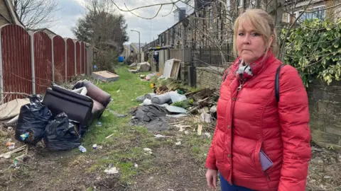 A woman wearing a red quilted jacket standing at the mouth of an alleyway filled with rubbish