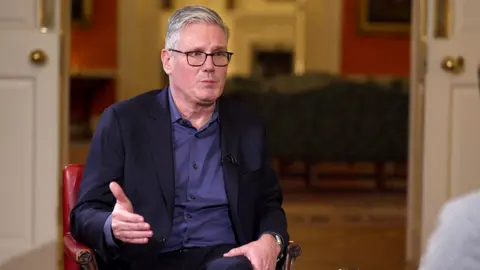 Sir Keir Starmer gestures while sitting in a red chair wearing a blue shirt and dark blazer