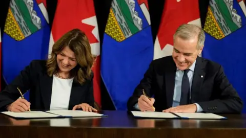 Canadian Prime Minister Mark Carney and Alberta Premier Daniel Smith sign a Memorandum of Understanding (MOU) ahead of the Prime Minister's Statement on Energy in Calgary, Alberta, Canada, November 27, 2025. They sit and pretend to each sign a document. Behind them is a row of Canadian and Alberta flags. 