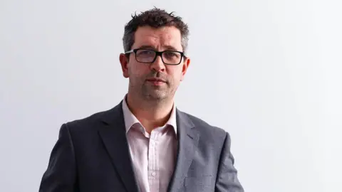 GWR A professional headshot of Dan Panes. He stands against a white backdrop and wears a grey suit jacket and a white shirt. He has short dark brown hair and wears black rimmed glasses.