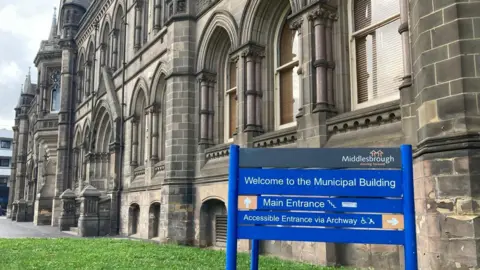 Daniel Hodgson The ornate gothic brick building housing Middlesbrough Council. It has arched windows and a sign in front of it for Middlesbrough Council. There is a grassed area and walkways.